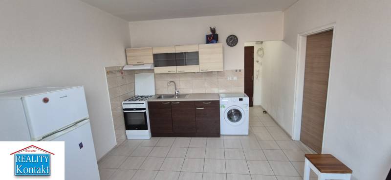 A kitchen in a studio apartment with a sink, stove, washing machine, and refrigerator on tiled flooring.