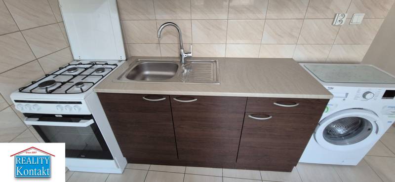 Kitchen in a studio apartment with a stove, sink, and washing machine, light tiled wall.