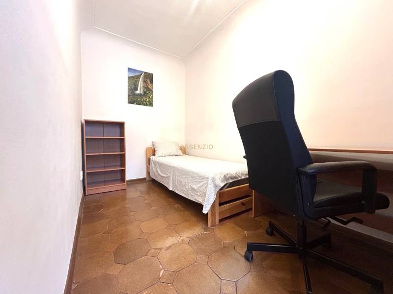 A room in a 3-room apartment with a bed, chair, shelf, and a picture on the wall.