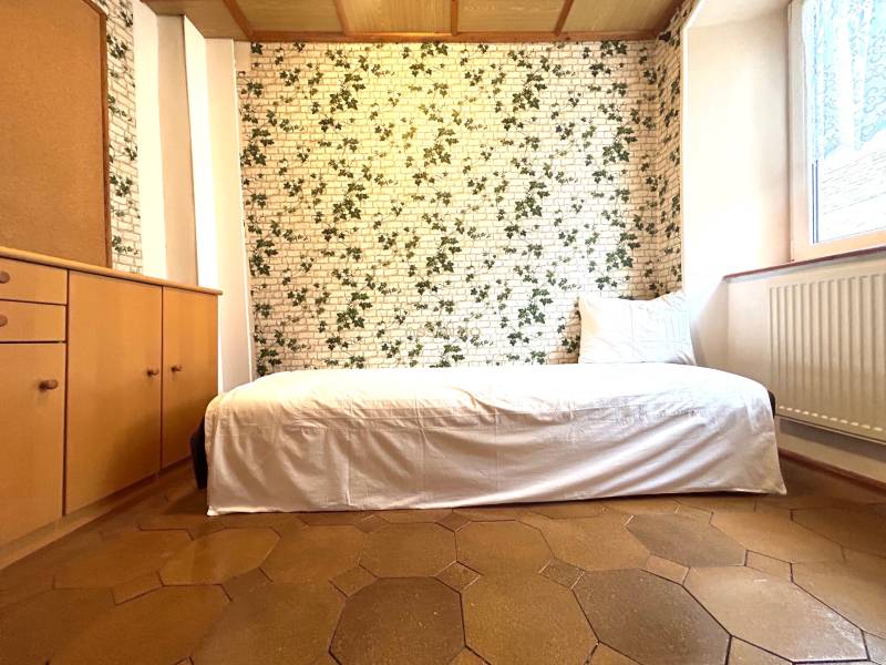 A room in a 3-room apartment with a single bed, patterned wallpaper, and wooden furniture.