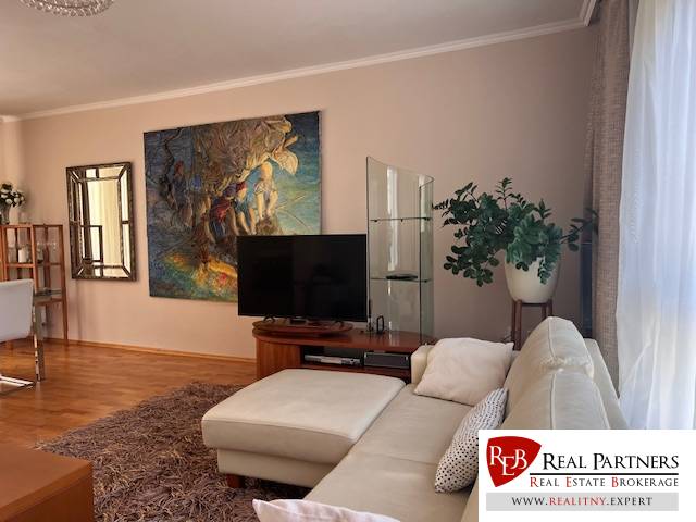 Living room in a 3-room apartment with a couch, television, plants, and a colorful painting on the wall.