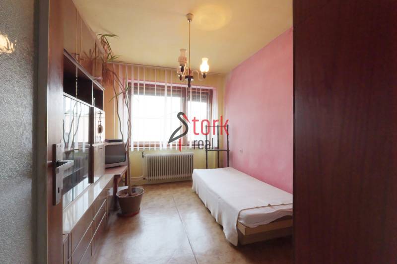 A room in a family house with a bed, cabinets, a plant, and a window with a radiator.