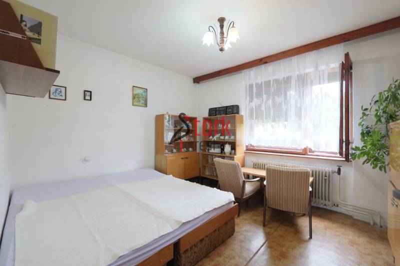 A room in a family house with a bed, a display cabinet, a table, and a plant by the window.