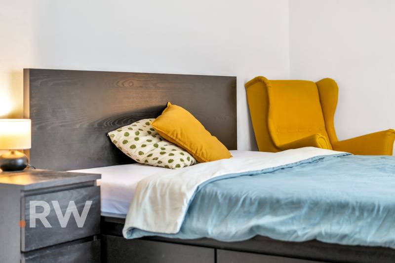 Bedroom with a bed and a nightstand in a 3-room apartment, yellow chair in the corner.