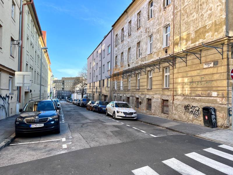 A narrow city street with parked cars, sidewalks, and older buildings.