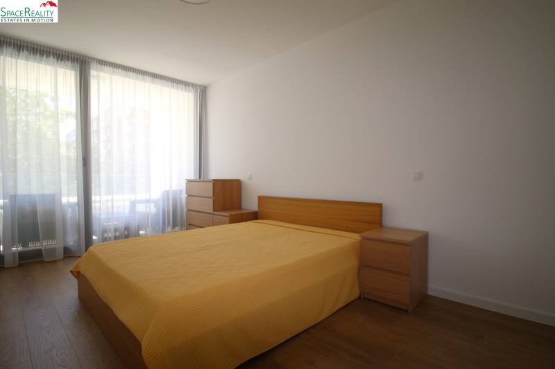 A bedroom with a bed and wooden furniture in a two-room apartment, light curtains letting in daylight.