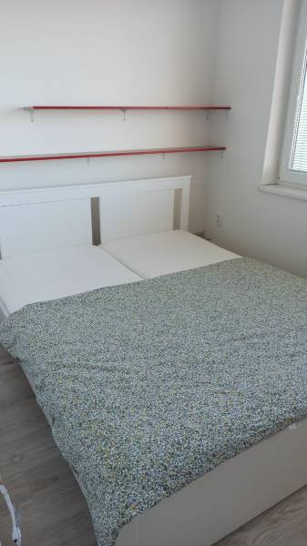 A bedroom with a double bed, white furniture, and two wall shelves in a two-room apartment.