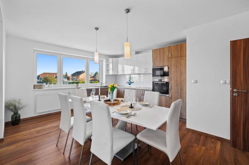 The kitchen on Pluhová Street, in a family house, contains white chairs, a wooden floor, large windows.