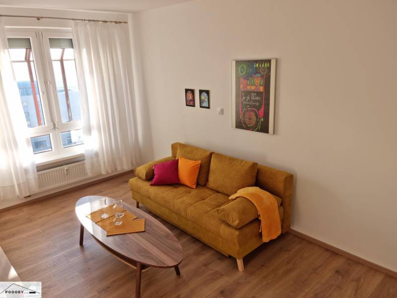 Living room of a two-room apartment with a yellow sofa, wooden table, and large window.