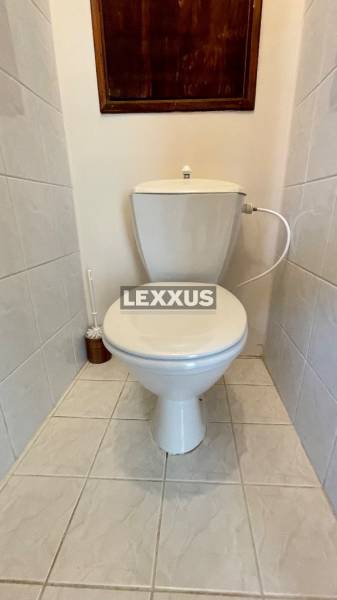 A toilet in a 3-room apartment with white tiles and a wooden panel on the wall.