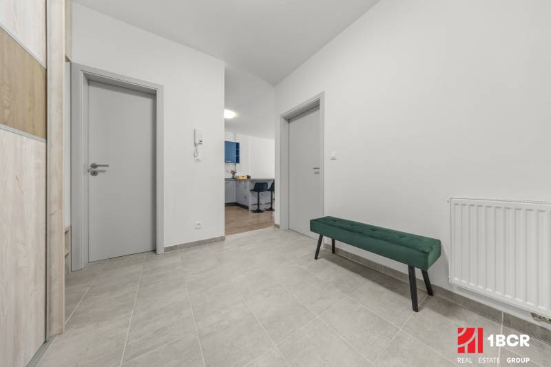 A hallway in a 2-room apartment with doors, a green bench, and a view into the kitchen.