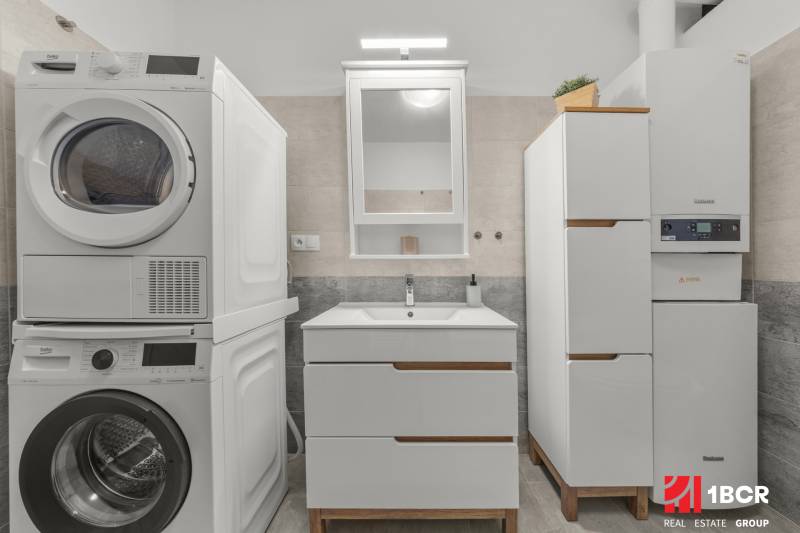 A bathroom in a 2-room apartment with a washing machine, dryer, sink, and mirror.