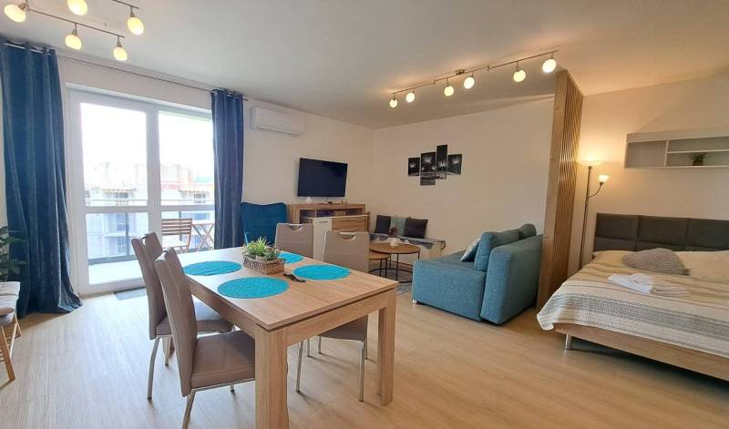 Studio apartment with a dining table, sofa, bed, and television, illuminated by lamps.