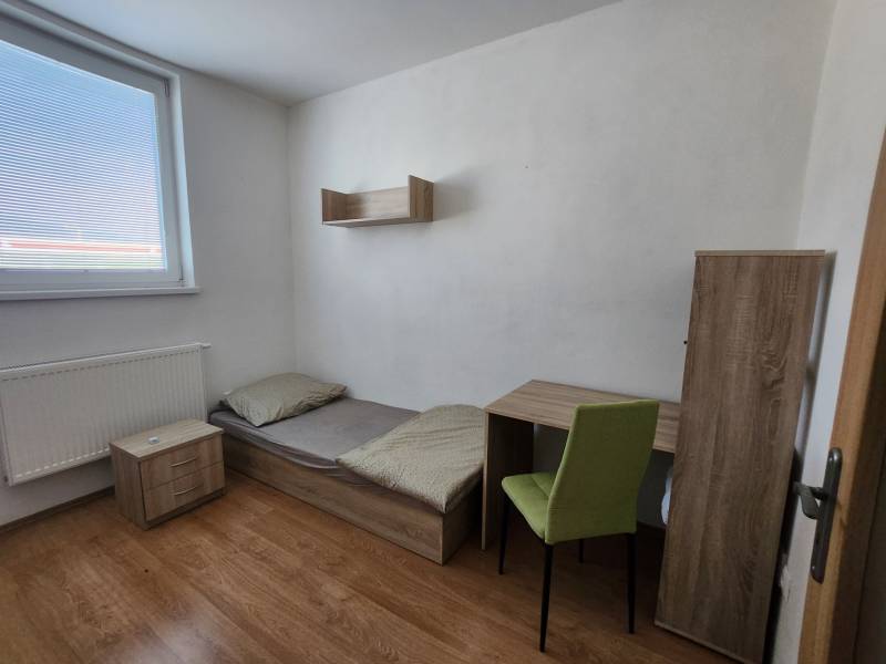 A simple room with a bed, a table, a chair, a nightstand, and a shelf in a building.