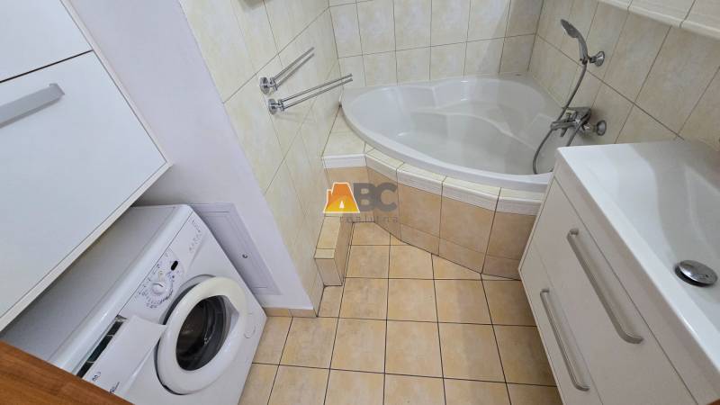 A bathroom with a corner bathtub, sink, and washing machine in a 2-room apartment.