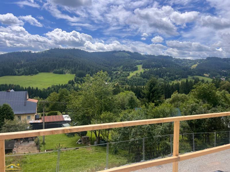 The view from the cottage in Korňa includes green meadows, hills, and a sky with clouds.