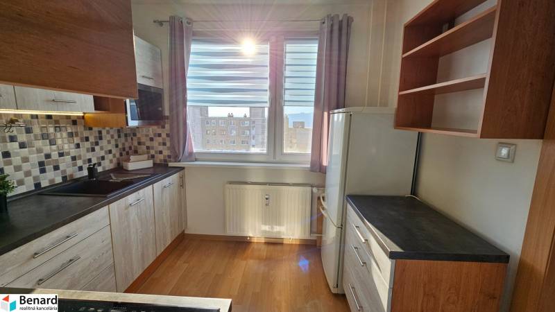 A kitchen in a studio apartment with wooden cabinets, tiles, and a window letting in light.