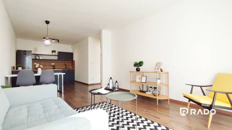 A living room in a two-room apartment with simple furniture, wooden flooring, and a kitchenette.