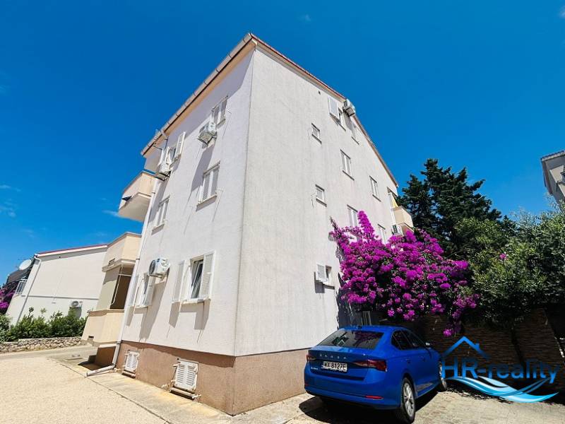 Apartment building in Novalja with parking, white-flowered trees, and a blue car in front of the building.