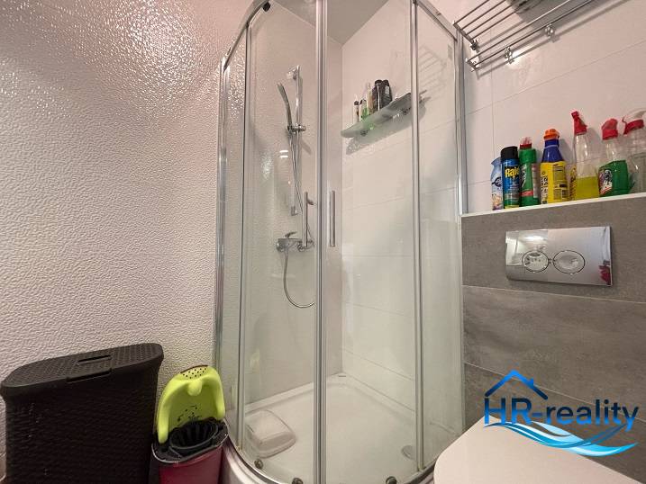 A bathroom in a 2-room apartment with a corner shower and a shelf for cleaning products.