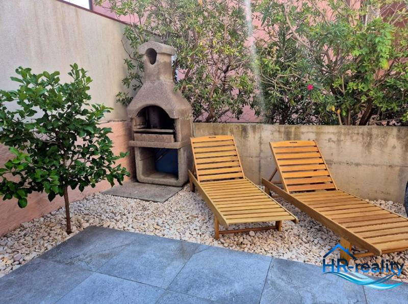 Garden space of a 2-bedroom apartment in Novalja with sunbeds and a grill surrounded by greenery.