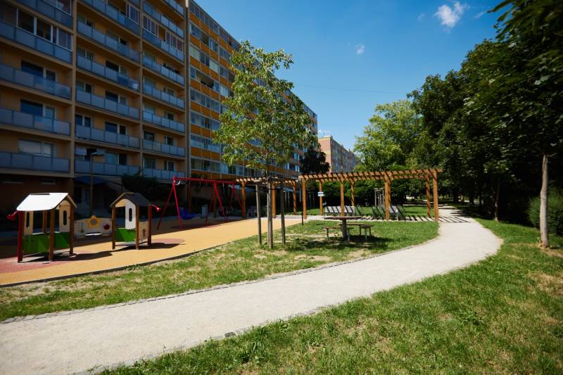 A playground with an apartment building in Košice - Juh district, surrounded by greenery and a walkway.