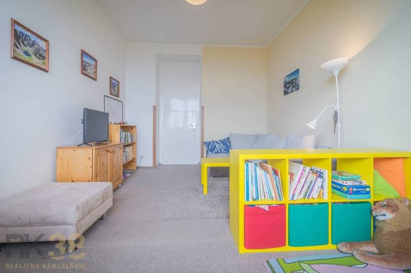A room in a 3-room apartment contains a sofa, a television, shelves with books, and colorful boxes.