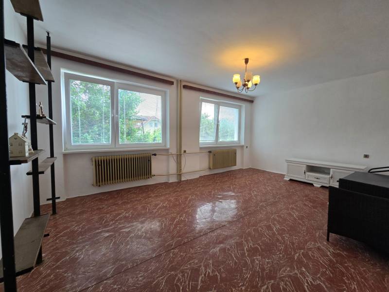 Living room in a family house, with white walls and a red floor, two windows, and furniture.