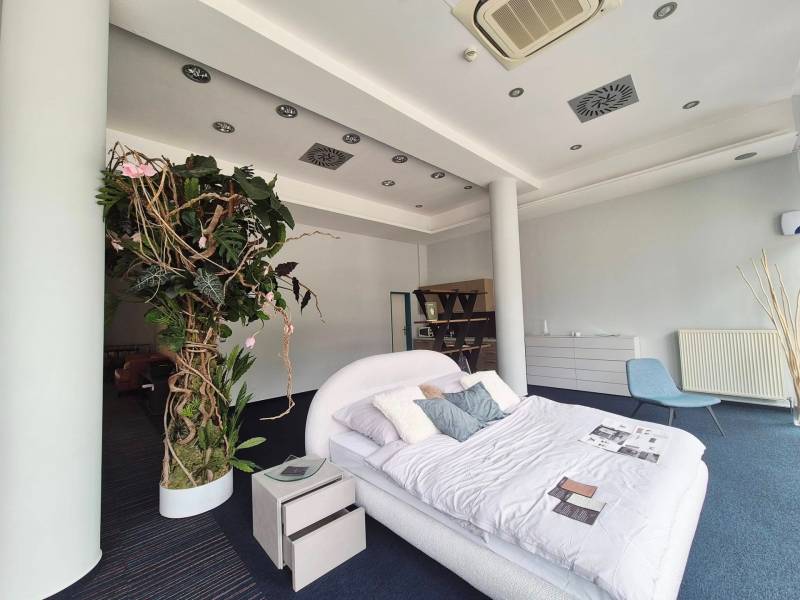 A bed with pillows and plants in commercial premises, next to a nightstand and a chair.