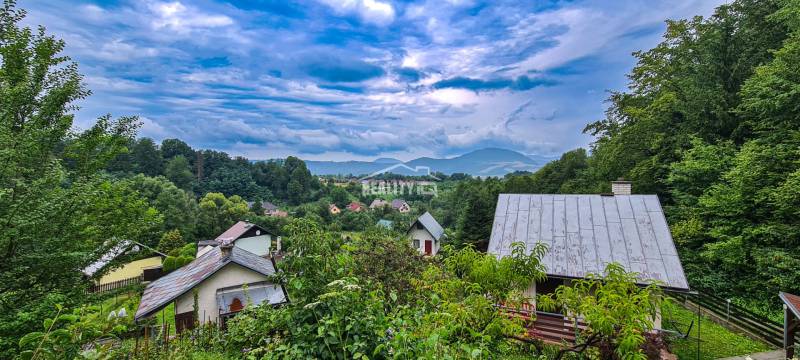 View of cottages in Mojš surrounded by lush greenery and hills under a dramatic sky.