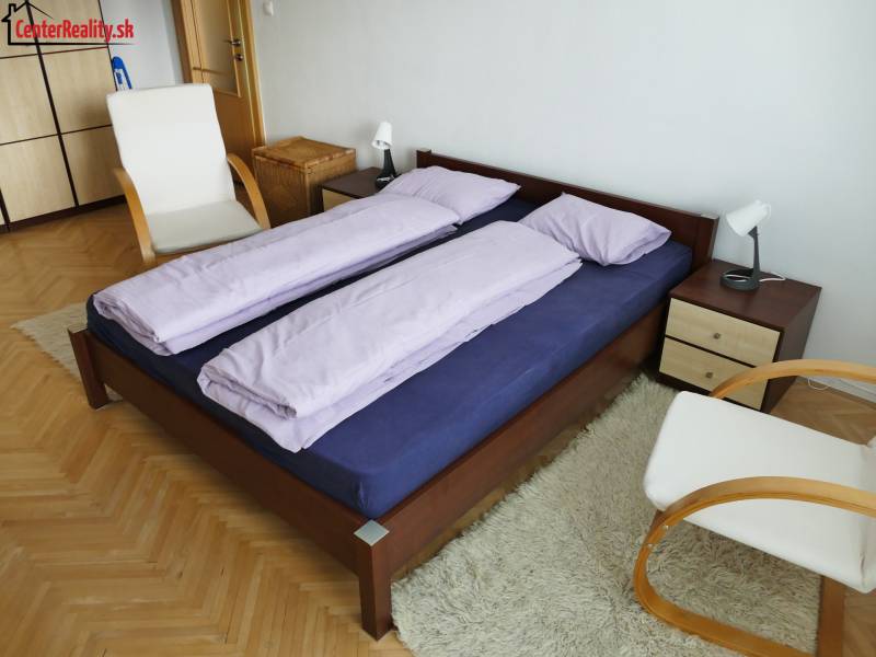 A bedroom in a 3-room apartment with a wooden bed, a chair, and a white carpet.