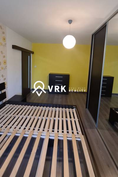 A bedroom in a 2-room apartment with a bed, two dressers, and a yellow wall.