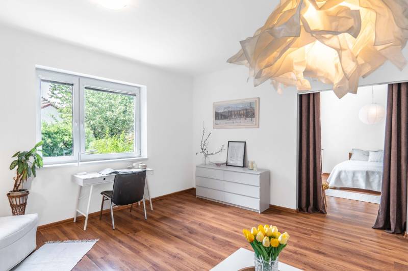 Bright living room with wooden flooring, a desk by the window, and a bed behind a curtain.