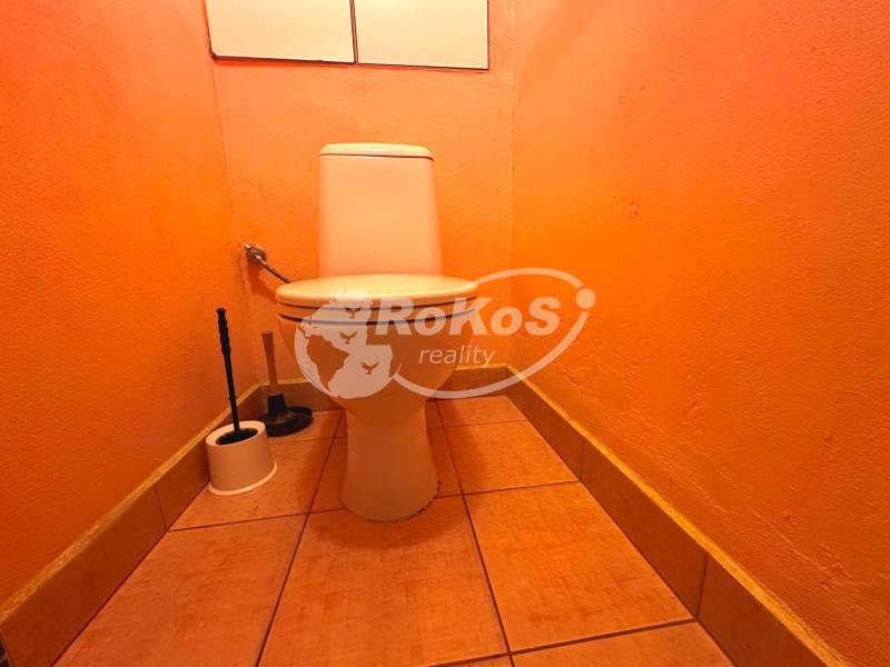 An orange toilet in a 3-room apartment with a toilet brush in the corner of the room.