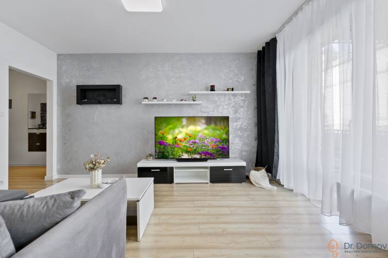 Living room in a 2-room apartment with a television, gray wall, and wooden floor.
