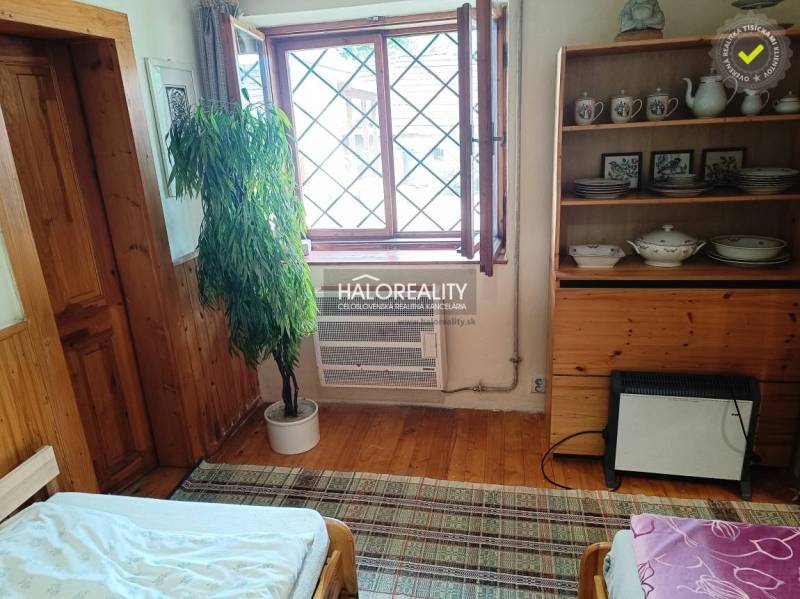 The interior of a cottage with wooden furniture, a plant, an open window, and a shelf with porcelain.