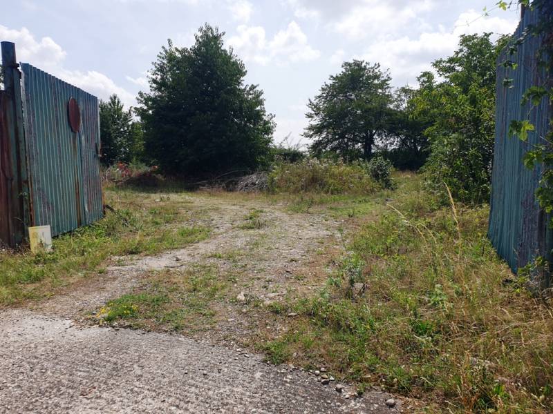 Fenced entrance to commercial land in Trenčianske Bohuslavice with greenery and a sidewalk.