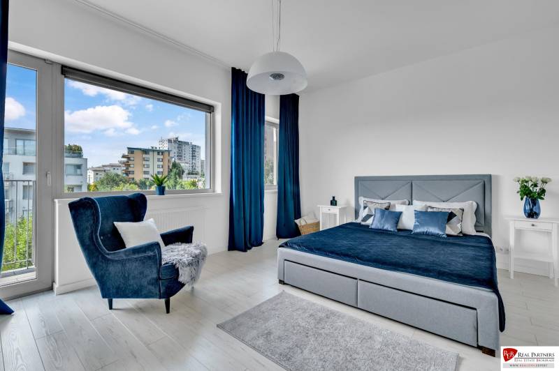 A bright bedroom in a 4-room apartment with a large window, blue curtains, and a comfortable chair.