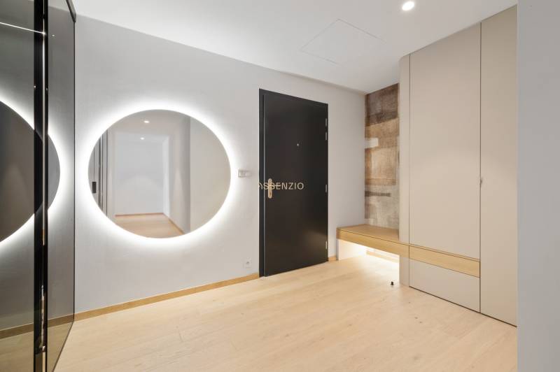 A hallway in a 2-room apartment with a lighted mirror, wooden floor, and storage space.
