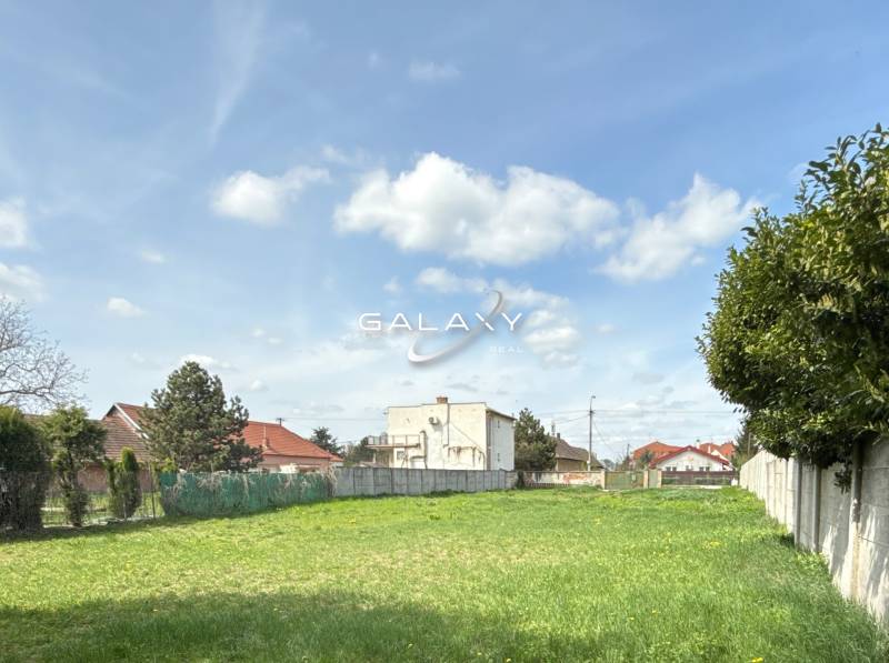 Land for residential use in Sládkovičovo, grass, fences, trees, buildings in the background, cloudy sky.