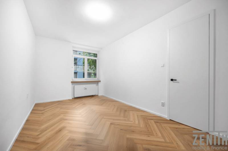 A room in a 3-room apartment with wooden flooring, white walls, and a window.