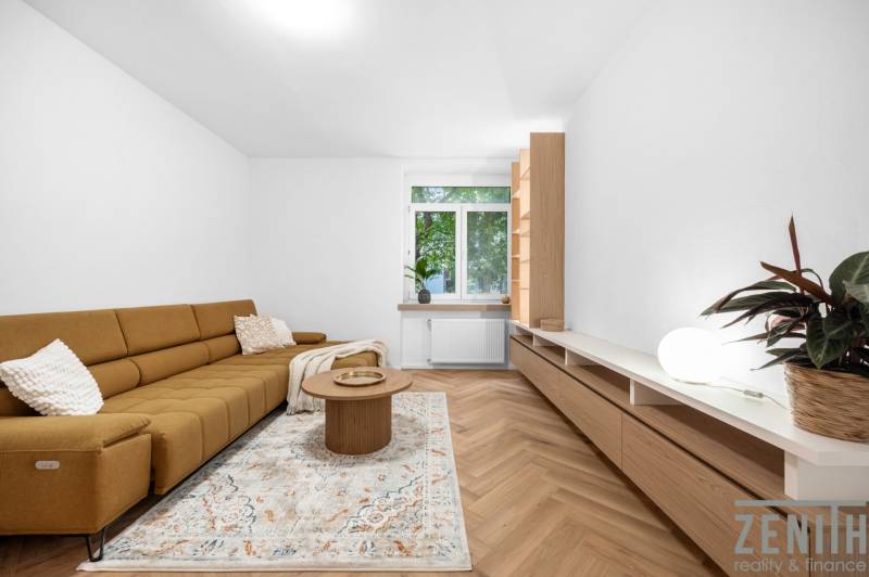 Living room in a three-room apartment with a brown sofa, a table, and a large window.
