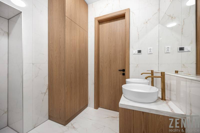 A bathroom with two sinks, wooden cabinets, and marble tiles in a three-room apartment.