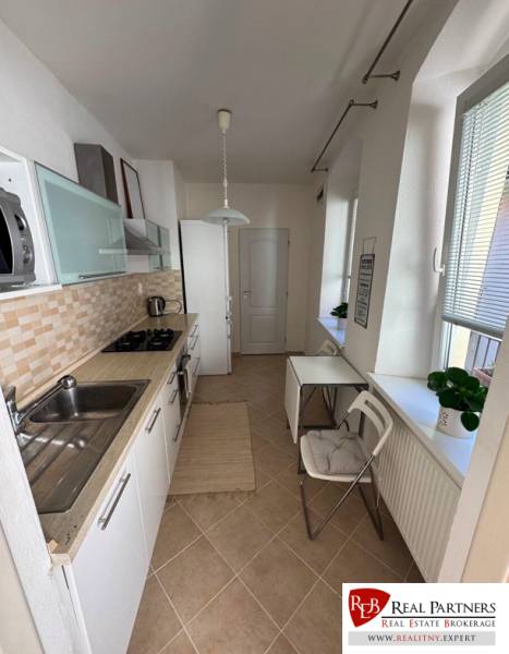 A kitchen in a 2-room apartment with white cabinets and windows with blinds.