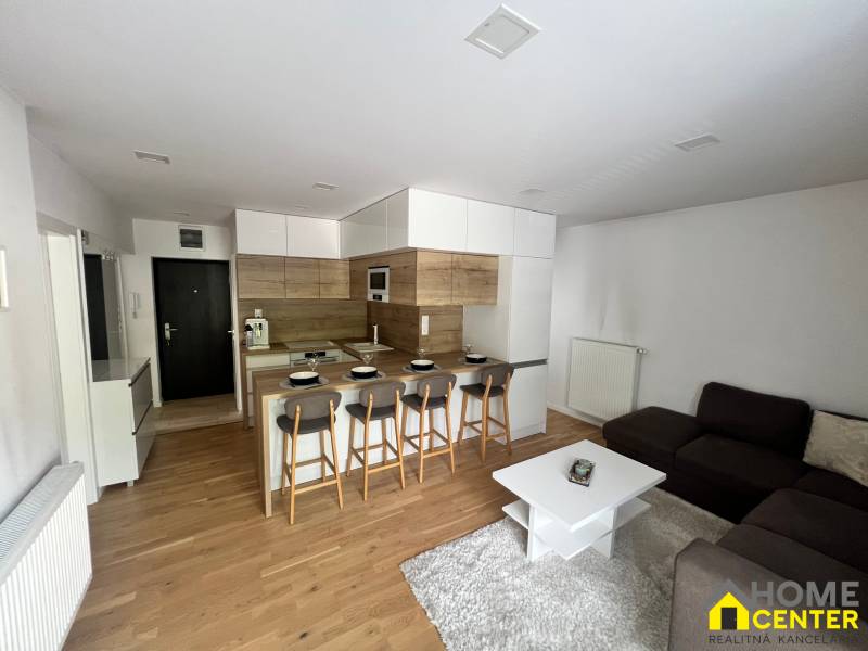 Living area of a 3-room apartment with an open kitchen, bar counter, and gray sofa.