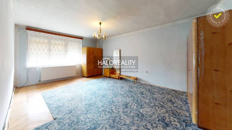 The interior of a cabin with a wooden wall, a tiled stove, and simple furniture in a cozy room.