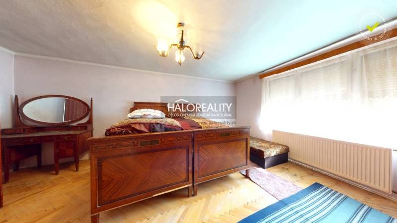 The interior of a cabin with a wooden bed, a tiled stove, and a carpet.