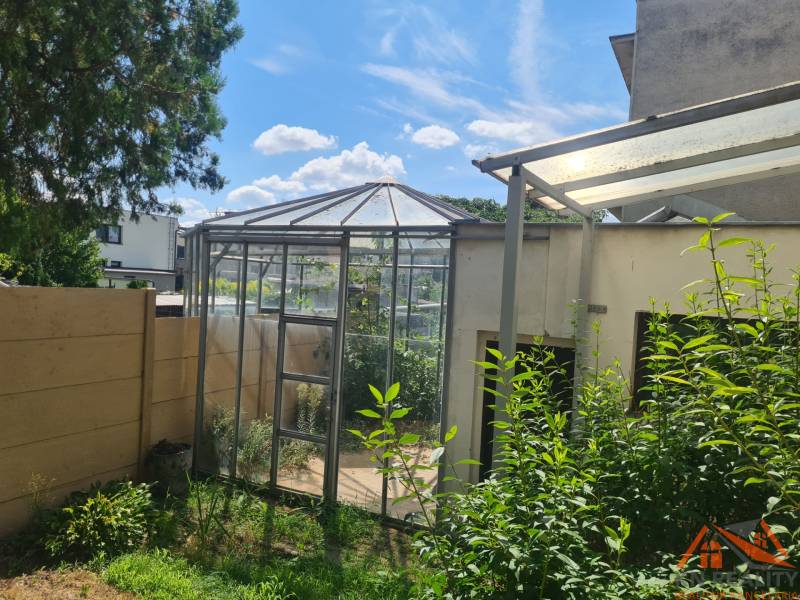 A greenhouse near a family house in Nové Zámky, surrounded by greenery and a fence.