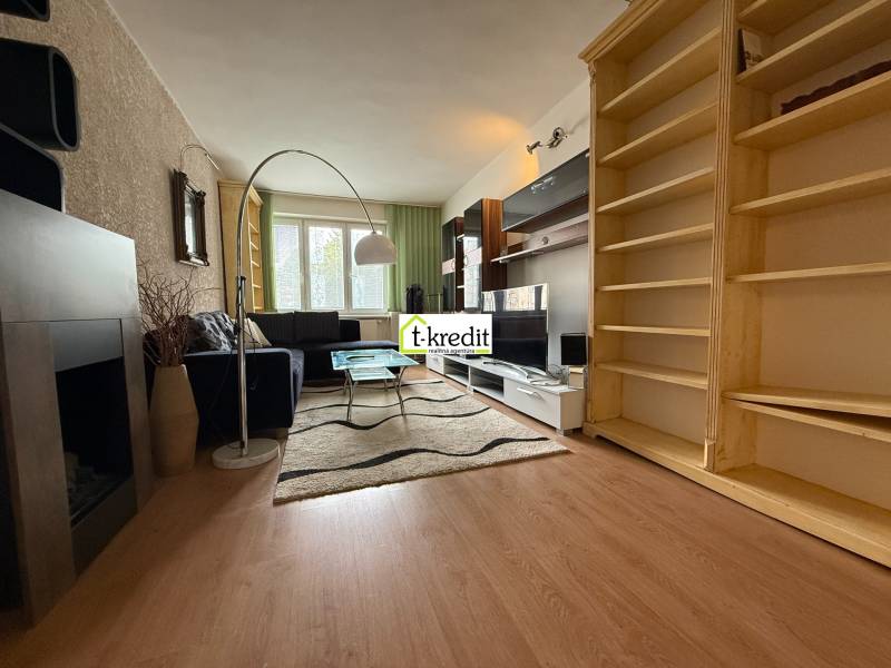Living room of a two-room apartment with a black sofa, carpet, and wooden shelves.