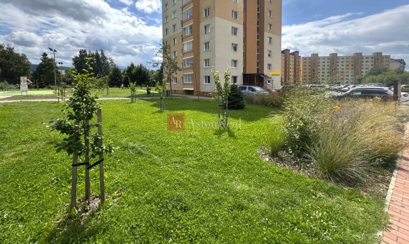 The grassy area in front of the apartment building on Západ Street in Tvrdošín, surrounded by greenery and a parking lot.